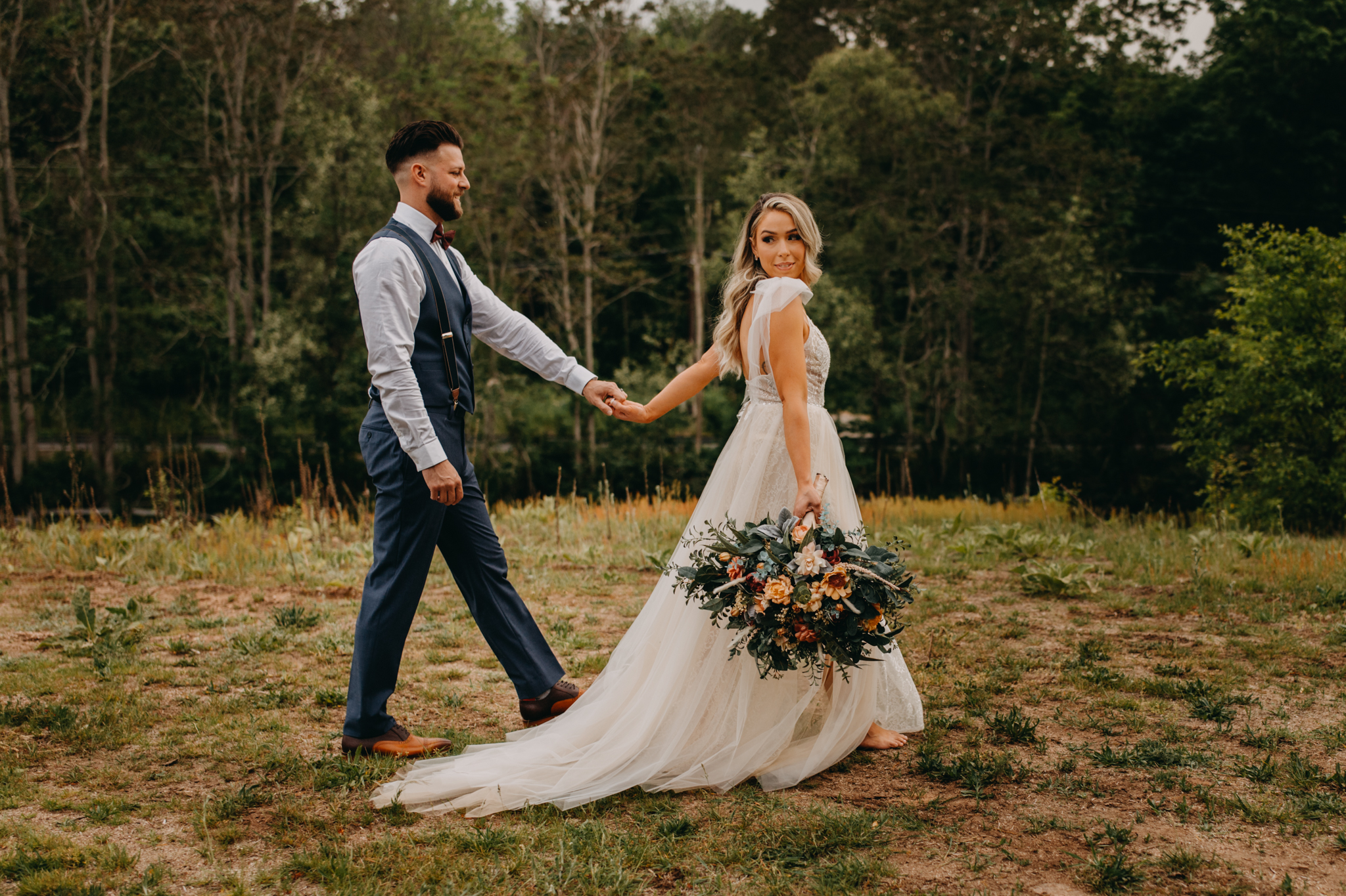 Bride and groom holding hands walking across a hill with a large and colorful bouquet at the Hinterland CT
