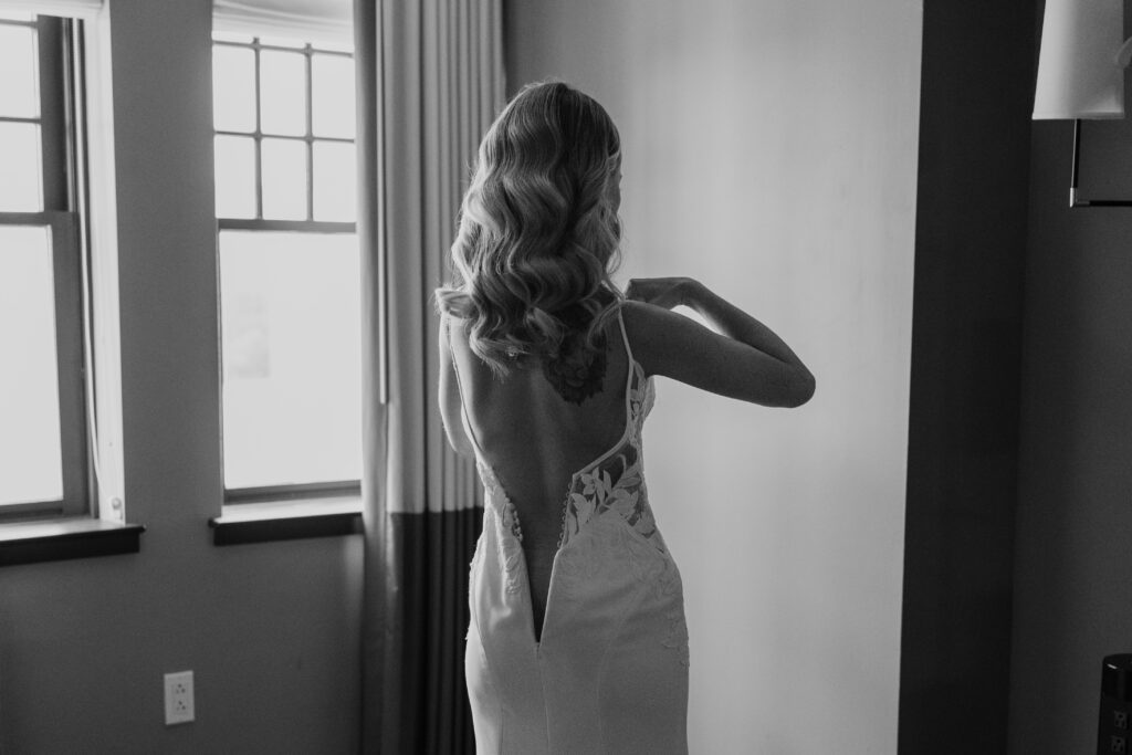 Bride is facing the window with back towards the camera adjusting the straps of her wedding dress