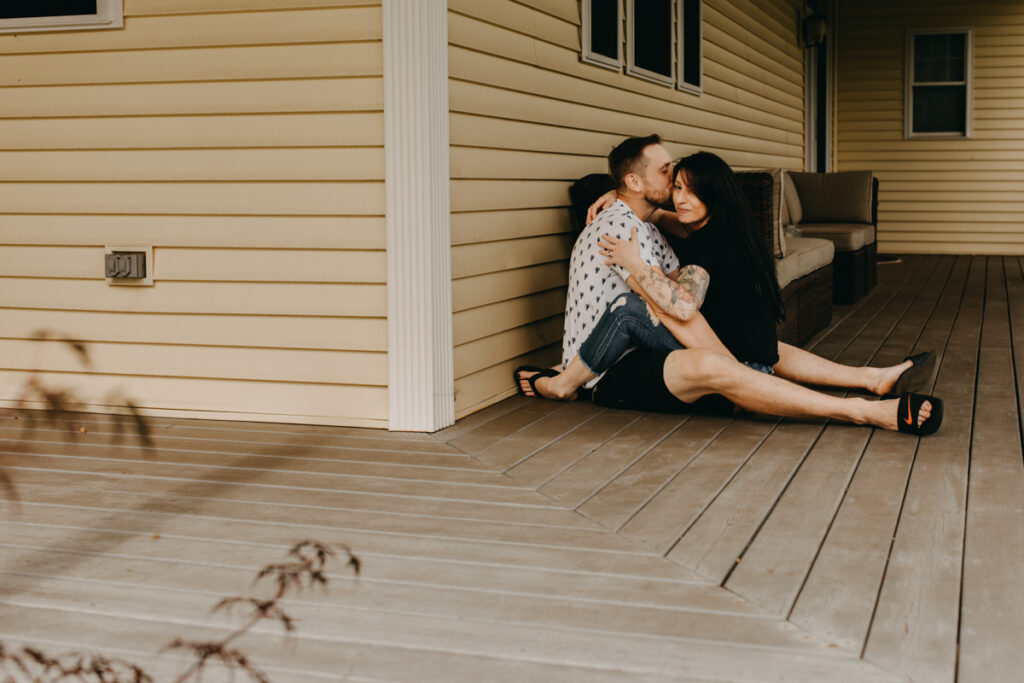 Man and woman sit facing each other with legs intertwined. Man kissing woman on temple, woman looking towards the camera smiling for in home photography session